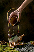 Hand pouring water into craft handmade ceramic teapot kettle, cup of hot green tea on moss with dry branch and leaves