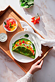 Crop person with spinach creppe and tomato and pepper slices and sour cream on tray
