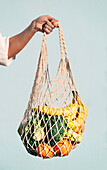 Cropped unrecognizable female standing with assorted fruits and vegetables in eco friendly mesh bag against blue wall in city