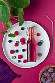 Glass bottle of colorful fruits juice served on plate with ripe berries on pink background