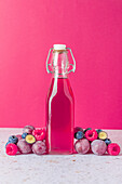 Glass bottle of fresh fruits juice surrounded with ripe berries served on table with glasses on pink background