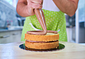 Crop female baker in apron standing and pouring chocolate cream on biscuit cake in light kitchen