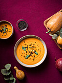 Bowl with delicious pumpkin soup served with seasonings placed near fresh pumpkin on purple table