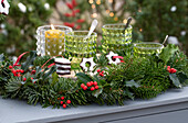 Glasses and lantern surrounded by a garland of fir branches and holly 'Blue Princess'.