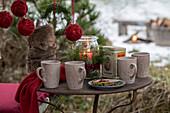 Mugs, lantern, and thermos bottle wrapped in fur on vintage garden table, Christmas tree ornaments wrapped in jute twine above it