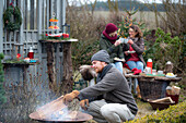 Man puts log in fire bowl, in the background two women toasting with mugs