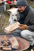 Mann mit Hund legt Holzscheit in die Feuerschale