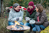 Three women with mulled wine squatting around fire pit