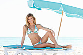 Junge blonde Frau im Bikini mit Palmen-Print auf einer Liege am Meer