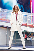 A long-haired woman wearing a white coat and trousers in front of a glass wall on the street