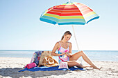 Young blonde woman in a bathing suit applying sunscreen under a parasol on the beach