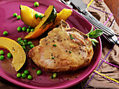 A pan fried bone-in pork chop with pan drippings sauce, slices of acorn squash and peas