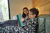 Mother watching son in pajamas reading book on sofa
