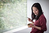 Young woman eating from bowl with chopsticks at window