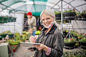 Happy female garden shop owner with clipboard in greenhouse
