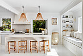 White kitchen with wooden stools around kitchen island, rattan pendant lights and built-in bench in front of window