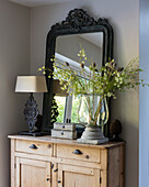 Framed mirror on wooden dresser with table lamp and large glass vase