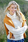 Junge blonde Frau in weißem Rollkragenpullover, mit gelbem Strickpulli über den Schultern in der Natur