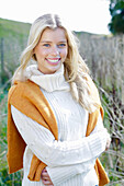 Young blond woman in white turtleneck sweater, with yellow knitted sweater slung over shoulders in nature