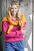Young blonde woman in a pink sweater with a yellow knit sweater over her shoulders in front of a wooden wall