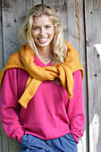 Junge blonde Frau in pinkfarbenem Pullover, mit gelbem Strickpulli über den Schultern vor Bretterwand