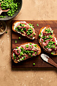 A pea, prosciutto, and ricotta crostini with a bite missing on a plate next to a bowl of peas, walnuts, and flakey sea salt