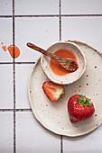Plate with ripe strawberries and bowl of sweet jam with spoon placed on tiled table