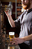 Cropped male barkeeper with red hair pouring beer on tap into glass while standing at counter during work in bar