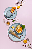 Unpeeled oranges on bowl with waffle cone on plate with almond and flower petals arranged with spoon peel and cup in light purple background