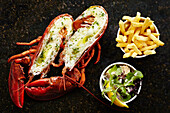 Cooked lobster split into two, in shell with garlic and herb butter, crispy fries, side salade with lettuce, lemon, feta, pumkin seeds