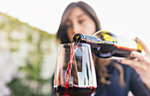 Cropped unrecognizable ethnic woman with long hair filling goblet with red wine from bottle with blank label during degustation session in restaurant