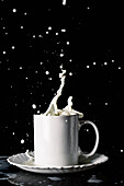 Ceramic mug on plate with flying splashes of milk on table against black background