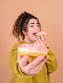 Cheerful young female eating appetizing sweet doughnut and giving to camera in studio against beige background
