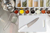 Top view of cutting board with knife placed near assorted sauces and flavoring in light kitchen with metal basket and bowl