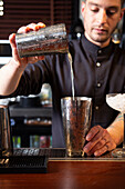 Crop concentrated young male bartender mixing drinks in shaker while preparing delicious cocktail at counter in restaurant