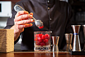 Crop professional male bartender in black uniform putting fresh raspberries into glass while preparing cocktail at counter in bar
