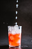 Ice cubes pouring into glass with alcoholic red cocktail with drops served on gray table against dark background in studio