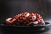 Tasty octopus tentacles served on dark plate on table against gray background in light kitchen during cooking process
