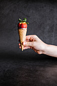 Hand of crop unrecognizable person holding tasty sweet ice cream cone with ripe red strawberry against gray background in studio