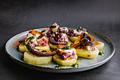 Tasty octopus tentacles on potato slices served on round ceramic plate on gray background in light kitchen