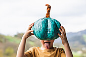 Lustiges Kind mit ausgestreckter Zunge hält türkisfarbenen Kürbis vor sein Gesicht