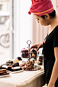 Hispanic woman in headscarf arranging desserts on table near window while working in bakery in daytime