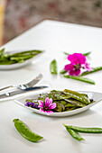 Delicious roasted snow peas garnished with flowers and served on plate near silverware on white table during lunch