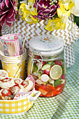 Vegetables in storage jar, wooden cutlery, mug, napkins and bouquet of flowers on picnic blanket