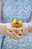 Person hält einen Muffin mit kleinen gelben Blüten als Dekoration