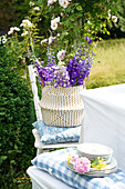 Rittersporn-Strauß in Vase auf Gartenstuhl mit Kissen, im Vordergrund Tellerstapel mit Rosen dekoriert auf Bank