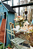 A bouquet of flowers and a nesting house on a table with a chair in front of it in a greenhouse