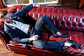 Young man with a beard in a jacket and jeans lies on a leather sofa
