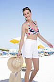 Junge brünette Frau in gestreiftem Bikinioberteil und weißen Shorts am Strand