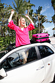 Young blond woman in a pink sundress stands in the skylight opening of a car